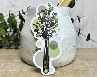 Puede incluir: Una pegatina con una ilustración estilizada de un tubo de ensayo lleno de flores y vegetación. Las flores tienen pétalos blancos y centros amarillos. El tubo de ensayo está rodeado de formas circulares verdes y grises. La pegatina está sobre un jarrón de cerámica moteado.
