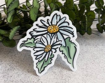 May include: A die-cut sticker featuring a hand-drawn illustration of two white daisies with yellow centers and green leaves. The artwork uses bold black outlines and a white border. The artist's signature is visible in the lower right corner.