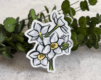 May include: A die-cut sticker featuring a hand-drawn illustration of three white daffodil flowers with yellow centers and green stems. The artwork uses bold black outlines and includes a small signature that reads PM21 near the top right flower.