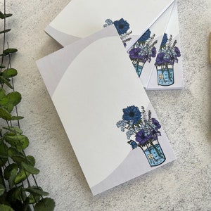 May include: A stack of white notepads with a purple and blue floral design in a glass beaker. The floral design is on the front of each notepad.