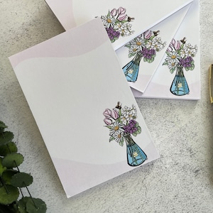 May include: A stack of white notepads with a pink gradient border. Each notepad features a hand-drawn illustration of a bouquet of flowers in a blue vase. The flowers include tulips, daisies, and violets.
