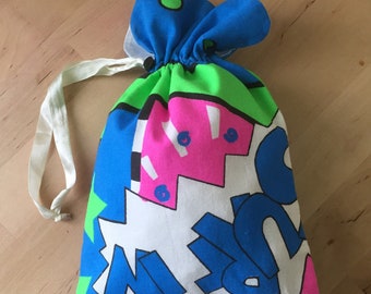 Op de afbeelding: Een kleurrijke trekkoordtas met een witte achtergrond en een geometrisch patroon in blauw, groen, roze en zwart. De tas heeft een wit trekkoord en is gemaakt van stof.