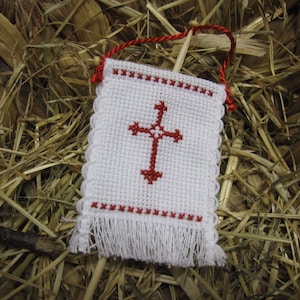 May include: A small white fabric banner with a red cross embroidered in the center. The banner has a white fringe around the edges and is hanging from a red string.