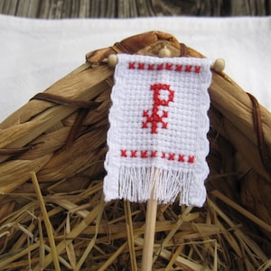 Puede incluir: Una pequeña bandera de tela blanca con una letra 'P' bordada en rojo sobre un palo de madera. La bandera está flecos y tiene un borde tejido. La bandera está sentada en una cesta tejida llena de paja.