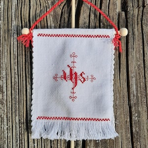 May include: A white, rectangular, cross-stitched wall hanging with red embroidered details. The design features a central cross and the letters IHS. It has a red rope hanger and fringe along the bottom edge. The background is a weathered wooden surface.