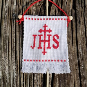 May include: A white cross-stitched banner with red lettering and a red cross design. The banner has a fringe at the bottom and is suspended by a red cord and wooden dowel. The background is a rustic wooden surface.