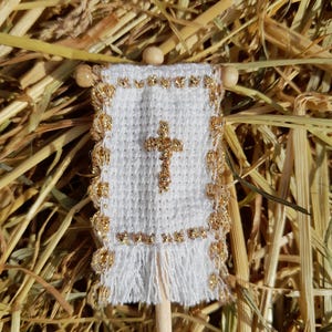 Può includere: Un piccolo stendardo bianco con una croce dorata e finiture dorate. Lo stendardo è attaccato a un tassello di legno con perline di legno nella parte superiore. Lo sfondo è paglia secca.