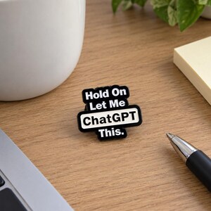 May include: A black and white enamel pin featuring the text "Hold On Let Me ChatGPT This." The pin has a rectangular design with rounded corners, white lettering on a black background, and a central white bar highlighting the word ChatGPT.