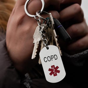 May include: A silver-toned metal keyring featuring a rectangular medical alert tag with the text COPD printed in black above a red Star of Life symbol. The tag is attached to a circular split ring along with several standard metal house keys.