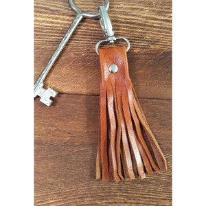May include: A brown leather tassel keychain with a silver clasp and rivet. The keychain is attached to a large, antique-style key. The leather is a warm, medium brown color, and the tassel is made of multiple strips of leather.