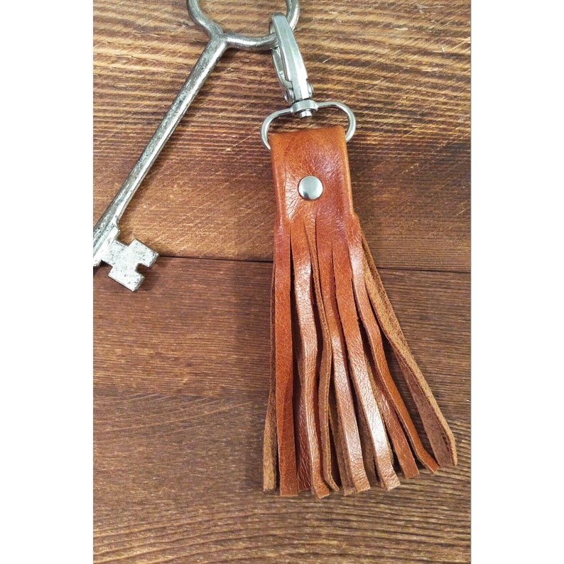 May include: A brown leather tassel keychain with a silver clasp and rivet. The keychain is attached to a large, antique-style key. The leather is a warm, medium brown color, and the tassel is made of multiple strips of leather.