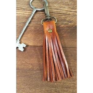 May include: A brown leather tassel keychain with a brass clasp and rivet. A vintage-style key is next to the keychain on a wooden surface. The tassel is made of multiple strips of leather.