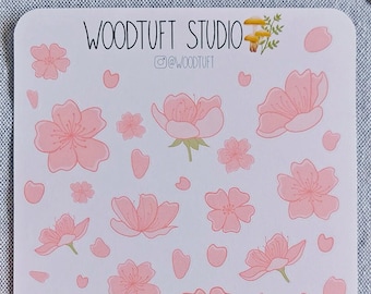 Pode incluir: Uma folha de adesivos de flores de cerejeira rosa e brancas com o texto "WOODTUFT STUDIO" e um pequeno ícone de câmera. Os adesivos estão em uma superfície cinza claro.