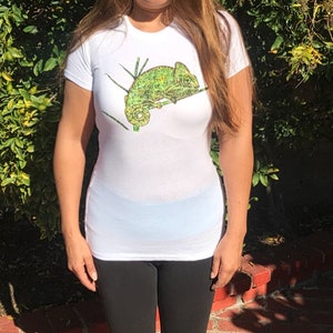 K&ouml;nnte beinhalten: Wei&szlig;es T-Shirt mit einem detaillierten Bild eines gr&uuml;nen Cham&auml;leons auf einem Ast. Das Cham&auml;leon ist in Gr&uuml;n- und Gelbt&ouml;nen dargestellt. Das T-Shirt hat einen Rundhalsausschnitt.
