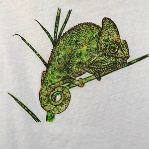 K&ouml;nnte beinhalten: Wei&szlig;es T-Shirt mit einer detaillierten Illustration eines gr&uuml;nen Cham&auml;leons auf einem Ast mit gr&uuml;nem Gras. Das Cham&auml;leon hat ein strukturiertes, gepunktetes Muster und ist im Profil.