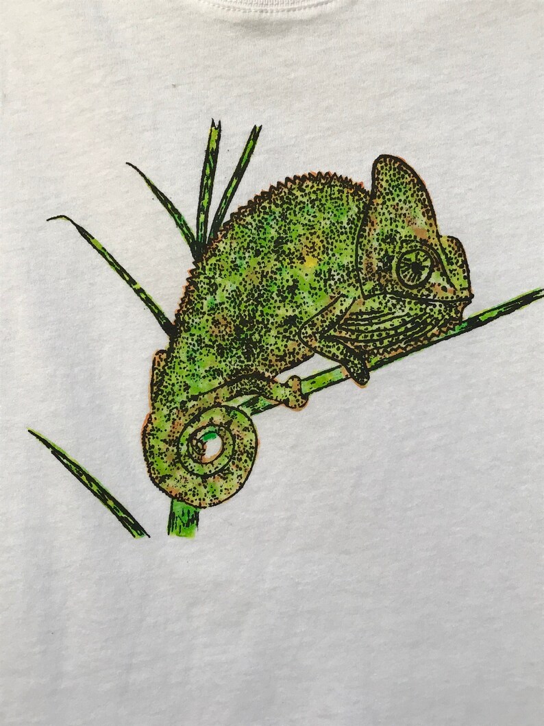 K&ouml;nnte beinhalten: Wei&szlig;es T-Shirt mit einer detaillierten Illustration eines gr&uuml;nen Cham&auml;leons auf einem Ast mit gr&uuml;nem Gras. Das Cham&auml;leon hat ein strukturiertes, gepunktetes Muster und ist im Profil.