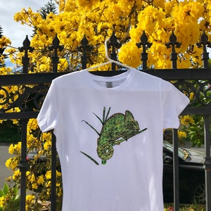 K&ouml;nnte beinhalten: Wei&szlig;es T-Shirt mit einem gr&uuml;nen und gelben Cham&auml;leon-Design. Das kurz&auml;rmelige Oberteil h&auml;ngt an einem wei&szlig;en Kleiderb&uuml;gel vor einem Hintergrund aus gelben Blumen und einem schwarzen schmiedeeisernen Zaun. Das Cham&auml;leon sitzt auf einem Ast.