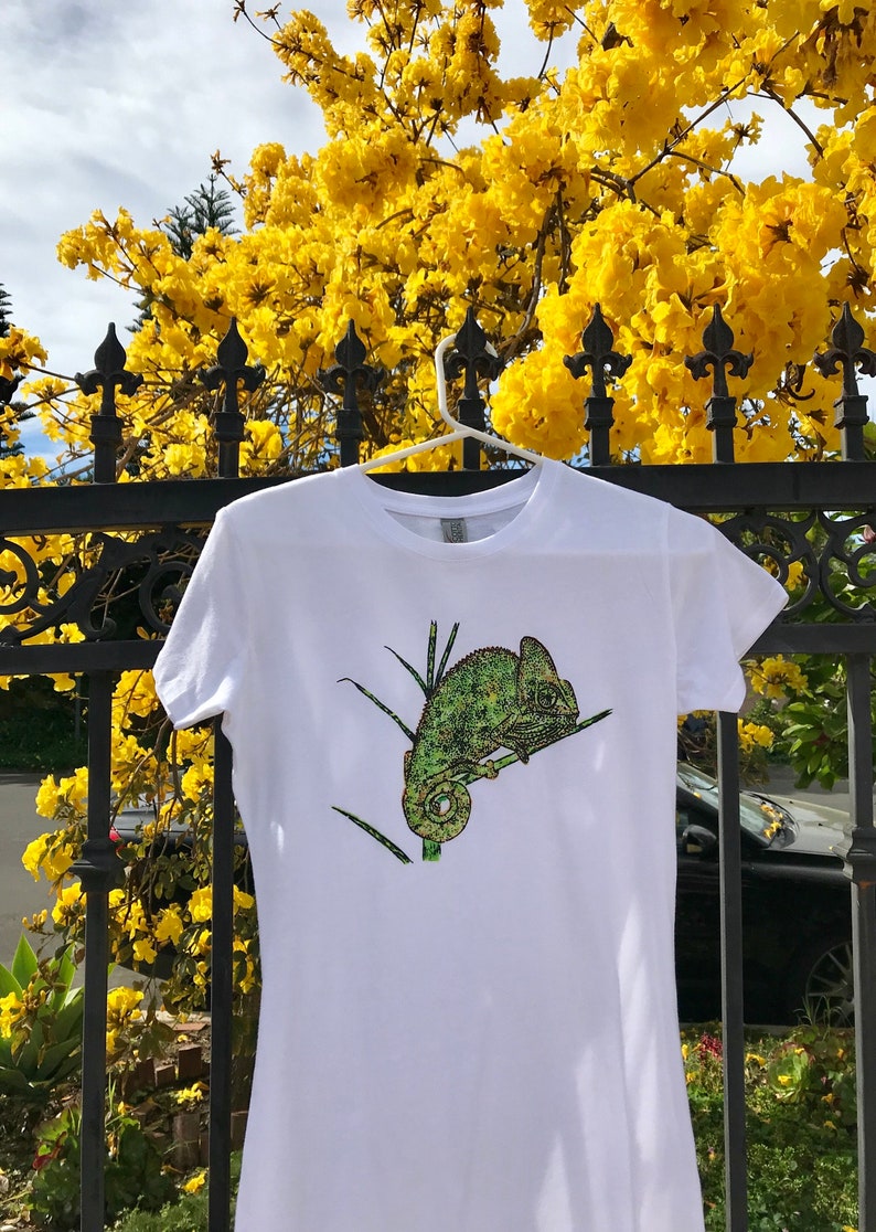 K&ouml;nnte beinhalten: Wei&szlig;es T-Shirt mit einem gr&uuml;nen und gelben Cham&auml;leon-Design. Das kurz&auml;rmelige Oberteil h&auml;ngt an einem wei&szlig;en Kleiderb&uuml;gel vor einem Hintergrund aus gelben Blumen und einem schwarzen schmiedeeisernen Zaun. Das Cham&auml;leon sitzt auf einem Ast.