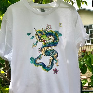 May include: White t-shirt with a blue and green dragon graphic. The dragon is surrounded by clouds and pink flowers. The shirt has a small tag with the Japanese character "力" on it.