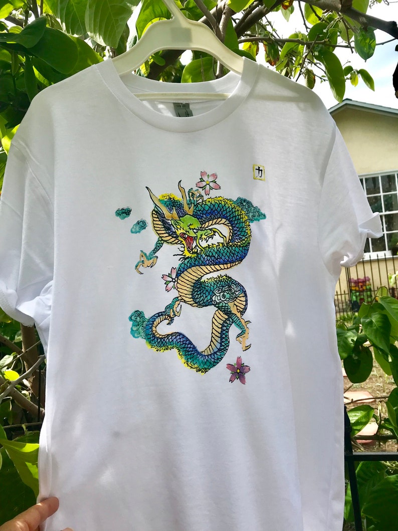 May include: White t-shirt with a blue and green dragon graphic. The dragon is surrounded by clouds and pink flowers. The shirt has a small tag with the Japanese character "力" on it.