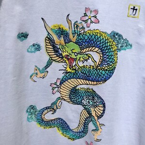May include: A white t-shirt with a colorful dragon design. The dragon is green, blue, and yellow with a red mouth. The dragon is surrounded by clouds and flowers. The word "力" is printed in a square in the top right corner of the shirt.