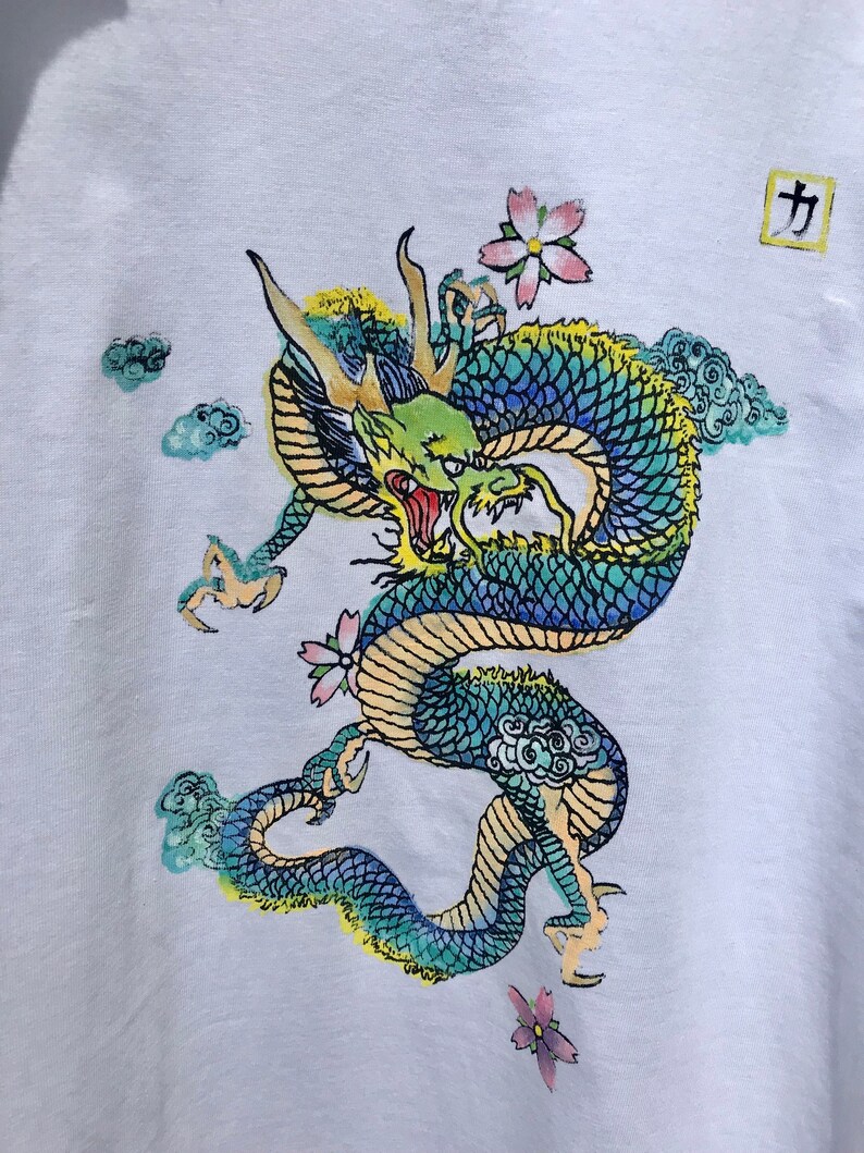 May include: A white t-shirt with a colorful dragon design. The dragon is green, blue, and yellow with a red mouth. The dragon is surrounded by clouds and flowers. The word "力" is printed in a square in the top right corner of the shirt.