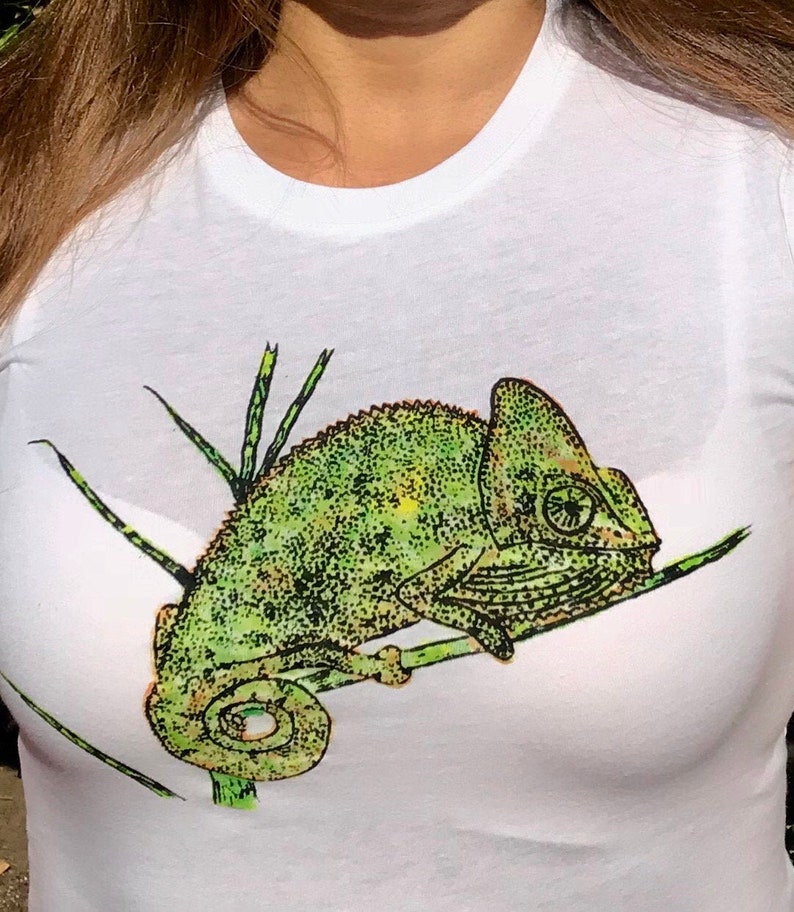 K&ouml;nnte beinhalten: Wei&szlig;es T-Shirt mit einem detaillierten gr&uuml;nen Cham&auml;leon-Motiv auf einem Ast. Das Cham&auml;leon hat ein strukturiertes Aussehen mit gelben und schwarzen Akzenten. Das Design ist mittig auf dem T-Shirt platziert.