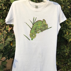 K&ouml;nnte beinhalten: Wei&szlig;es T-Shirt mit einer detaillierten Illustration eines gr&uuml;nen und gelben Cham&auml;leons auf einem Ast. Das Cham&auml;leon hat ein strukturiertes Aussehen. Das T-Shirt h&auml;ngt an einem wei&szlig;en Kleiderb&uuml;gel vor einem Hintergrund aus gr&uuml;nem Laub.