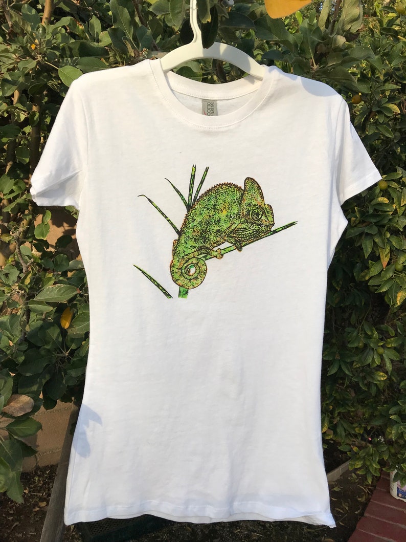 K&ouml;nnte beinhalten: Wei&szlig;es T-Shirt mit einer detaillierten Illustration eines gr&uuml;nen und gelben Cham&auml;leons auf einem Ast. Das Cham&auml;leon hat ein strukturiertes Aussehen. Das T-Shirt h&auml;ngt an einem wei&szlig;en Kleiderb&uuml;gel vor einem Hintergrund aus gr&uuml;nem Laub.