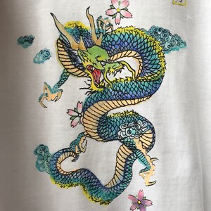 May include: A white t-shirt with a blue, green, and yellow dragon design. The dragon is surrounded by clouds and pink flowers. The Japanese character "力" is printed in the top right corner of the shirt.