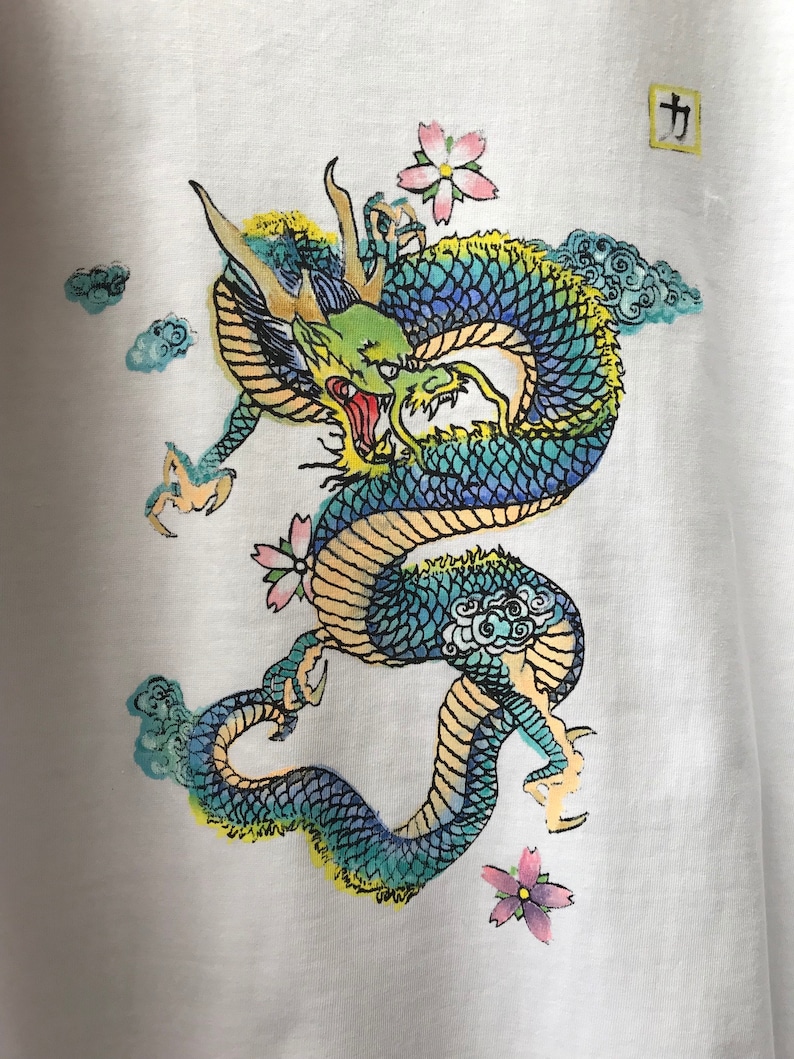 May include: A white t-shirt with a blue, green, and yellow dragon design. The dragon is surrounded by clouds and pink flowers. The Japanese character "力" is printed in the top right corner of the shirt.