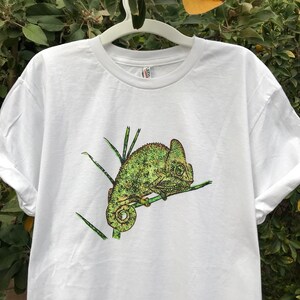 K&ouml;nnte beinhalten: Wei&szlig;es T-Shirt mit einer detaillierten Illustration eines gr&uuml;nen Cham&auml;leons auf einem Ast. Das Cham&auml;leon ist in Gr&uuml;n- und Gelbt&ouml;nen dargestellt. Das Design ist mittig auf dem T-Shirt platziert.