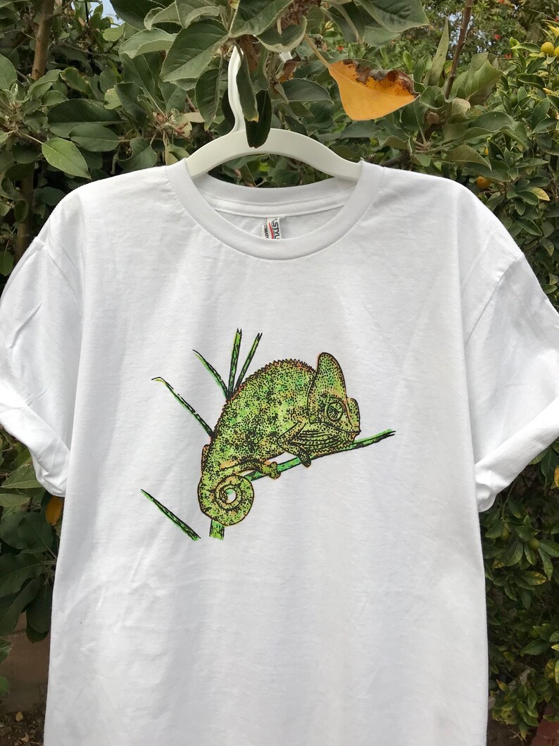 K&ouml;nnte beinhalten: Wei&szlig;es T-Shirt mit einer detaillierten Illustration eines gr&uuml;nen Cham&auml;leons auf einem Ast. Das Cham&auml;leon ist in Gr&uuml;n- und Gelbt&ouml;nen dargestellt. Das Design ist mittig auf dem T-Shirt platziert.