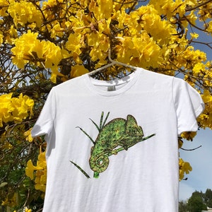 K&ouml;nnte beinhalten: Wei&szlig;es T-Shirt mit einer detaillierten Illustration eines gr&uuml;nen Cham&auml;leons auf einem Ast. Das Cham&auml;leon ist in Gr&uuml;n- und Gelbt&ouml;nen dargestellt. Das T-Shirt h&auml;ngt an einem wei&szlig;en Kleiderb&uuml;gel vor einem Hintergrund aus gelben Blumen und blauem Himmel.