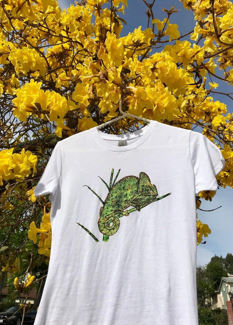 K&ouml;nnte beinhalten: Wei&szlig;es T-Shirt mit einer detaillierten Illustration eines gr&uuml;nen Cham&auml;leons auf einem Ast. Das Cham&auml;leon ist in Gr&uuml;n- und Gelbt&ouml;nen dargestellt. Das T-Shirt h&auml;ngt an einem wei&szlig;en Kleiderb&uuml;gel vor einem Hintergrund aus gelben Blumen und blauem Himmel.