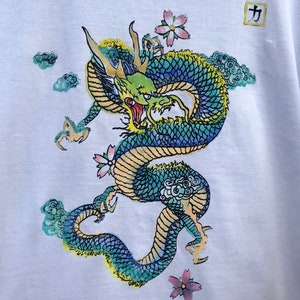 May include: A white t-shirt with a blue and green dragon design. The dragon is surrounded by clouds and pink flowers. The kanji character "力" (power) is printed in a square in the upper right corner of the shirt.