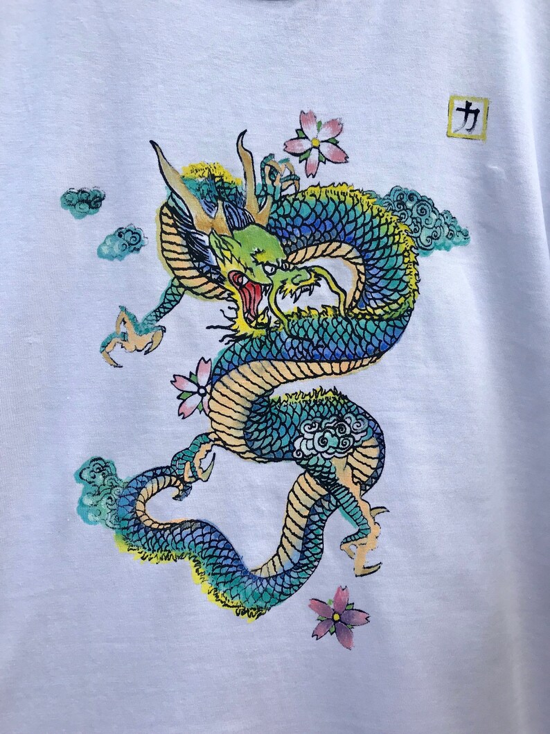 May include: A white t-shirt with a blue and green dragon design. The dragon is surrounded by clouds and pink flowers. The kanji character "力" (power) is printed in a square in the upper right corner of the shirt.