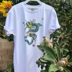 May include: White t-shirt with a blue and green dragon graphic and pink flowers. The dragon is curled up and has a fierce expression. The shirt has a crew neck and short sleeves.