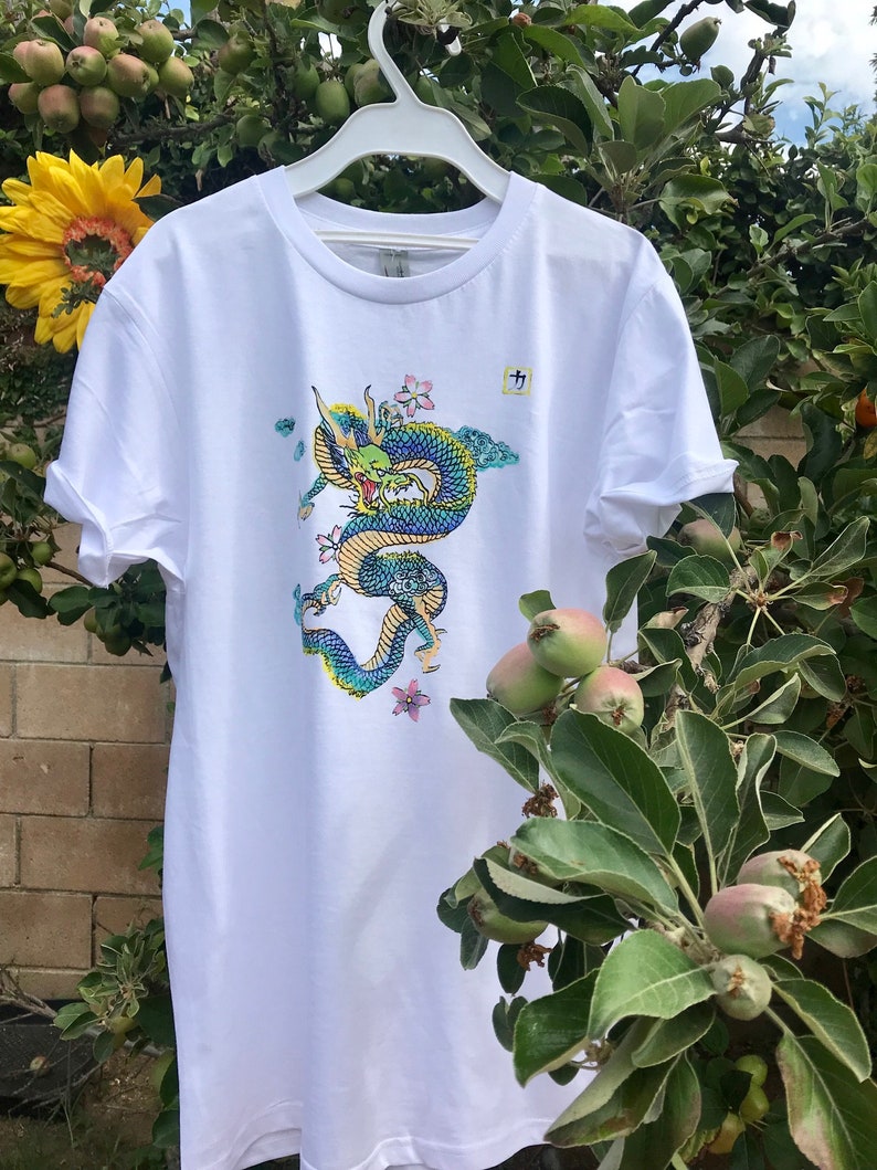 May include: White t-shirt with a blue and green dragon graphic and pink flowers. The dragon is curled up and has a fierce expression. The shirt has a crew neck and short sleeves.