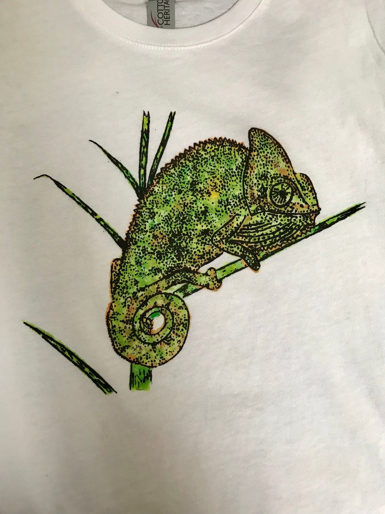 K&ouml;nnte beinhalten: Wei&szlig;es T-Shirt mit einer detaillierten Illustration eines gr&uuml;nen Cham&auml;leons auf einem Ast. Das Cham&auml;leon ist in einem punktartigen Stil dargestellt, mit Gr&uuml;n- und Braunt&ouml;nen. Das Design enth&auml;lt gr&uuml;ne, grasartige Elemente.