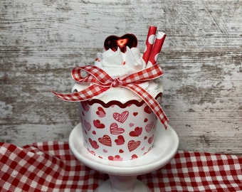 May include: A white cupcake with red and white polka dot straws and a red heart on top. The cupcake is decorated with a red and white gingham ribbon and is sitting on a white cake stand. The cake stand is on a red and white gingham tablecloth.