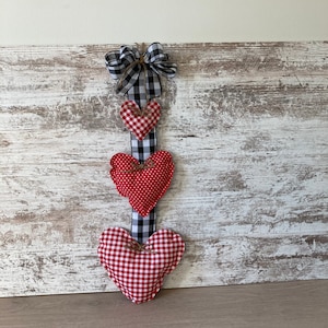 May include: A red and white checkered fabric heart garland with a black and white checkered ribbon. The garland is hanging on a whitewashed wood background.