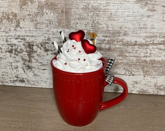 May include: Red mug with white whipped cream and red heart sprinkles topped with two red hearts and a small bear figurine. The mug has a black and white checkered ribbon tied around the handle.