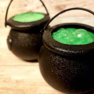 May include: Two black cauldron-shaped containers with green liquid inside. The cauldrons have handles and are made of a textured material.