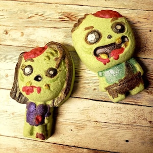 May include: Two green bath bombs shaped like zombies with red and white details. The zombie on the left is a female with brown hair and a purple shirt. The zombie on the right is a male with a green shirt.