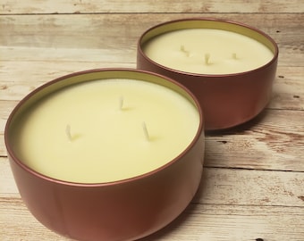 May include: Two rose gold candle jars with three wicks each. The candles are a pale yellow color and are sitting on a light wood surface.
