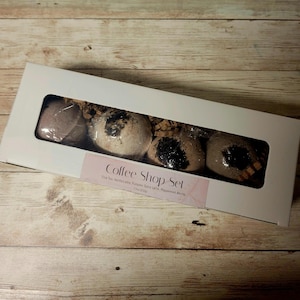 May include: A white rectangular box with a clear window displaying four coffee-themed bath bombs. The bath bombs are various shades of brown and tan, with dark accents. The box has a pink label that reads "Coffee Shop Set".