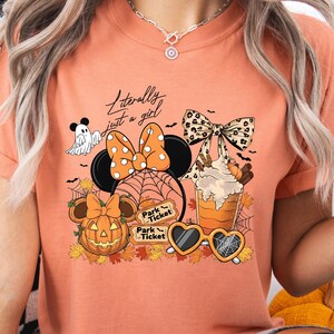 Op de afbeelding: Oranje T-shirt met een Halloween-thema. Het ontwerp bevat een Minnie Mouse-hoofd met een pompoen, spinnenweb en strik. De tekst "Literally just a girl" is ook aanwezig. Zonnebrillen en een spook zijn ook inbegrepen.
