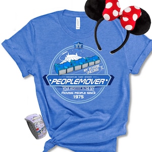 May include: A blue t-shirt featuring a graphic of the PeopleMover ride with the text "PEOPLEMOVER" and "YOUR HIGHWAY IN THE SKY MOVING PEOPLE SINCE 1975." The image also includes a pair of black Minnie Mouse ears with red polka dots and a purple and white watch.