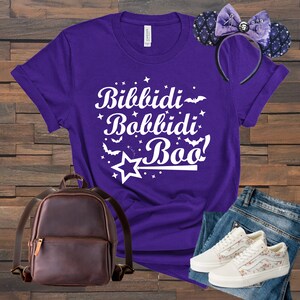 Puede incluir: Camiseta morada con texto blanco que dice "Bibbidi Bobbidi Boo!" con un gráfico de estrella y varita blanca. La camiseta se combina con una mochila marrón, jeans azules y zapatillas blancas con detalles florales.
